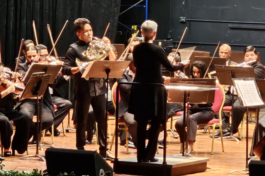 Nell Flanders stands on a stage in front of the Panama National symphony orchestra with guest artist Alberto Lin playing French horn and standing next to her